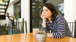 An adult feeling lonely while dating, sitting beside a window with a phone and a mug.